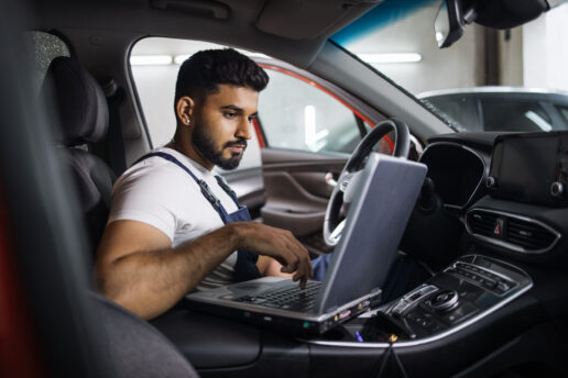 ADAS Calibration Young bearded man car technician mechanic sitting inside red car repairing problem of engine, during system checking detail, using laptop computer for maintenance and fixing in garage.